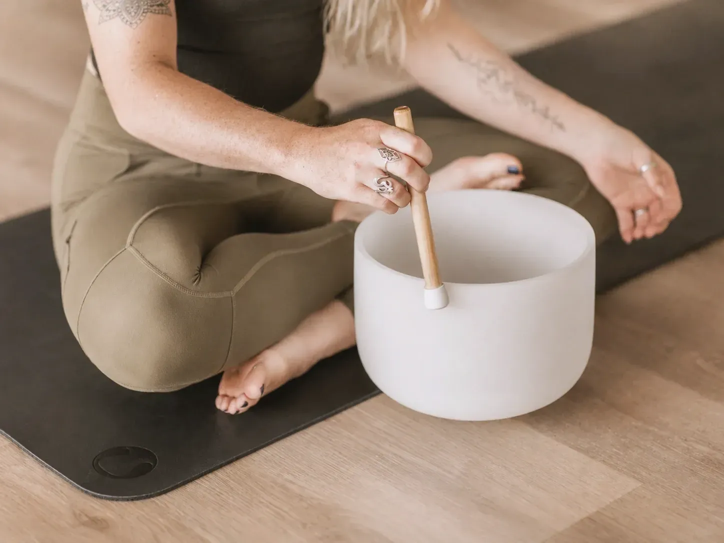 A woman seated cross-legged on her mat, sounding a singing bowl with a wooden striker